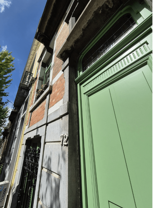 porte ancienne reproduction à l’identique Un bâtiment en brique haut et étroit avec une grande porte verte au premier plan. Il y a une fenêtre au deuxième étage avec un petit balcon. Un arbre est visible sur le côté gauche et le ciel est partiellement nuageux. Le numéro « 72 » est inscrit sur le mur à côté de la porte.
