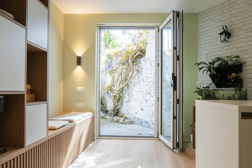 porte papillon Chambre lumineuse avec des touches de bois et des murs vert clair. Des portes vitrées ouvertes mènent à un patio avec un arbre torsadé contre un mur de briques blanches. Des plantes dintérieur sont posées sur un comptoir et un éclairage doux crée une atmosphère chaleureuse.
