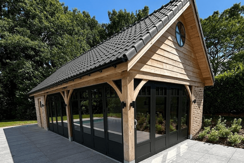 système accordéon Une structure moderne en bois avec un toit en tuiles incliné et de grandes portes vitrées se trouve sur un patio. Le bâtiment est entouré de verdure et darbres sous un ciel bleu clair. Une petite fenêtre circulaire se trouve près du sommet du toit.